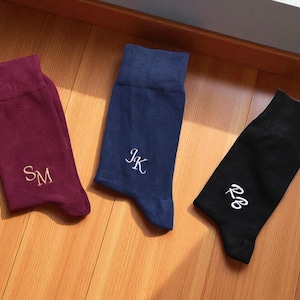 Può includere: Tre paia di calzini personalizzati in bordeaux, blu navy e nero. Ogni calzino presenta iniziali ricamate con filo bianco. I calzini sono disposti su una superficie di legno.