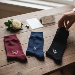 May include: Three pairs of crew socks in burgundy, navy blue, and black. Each sock features embroidered initials in a contrasting thread color: SM in gold on the burgundy sock, JK in white on the navy sock, and RB in white on the black sock.