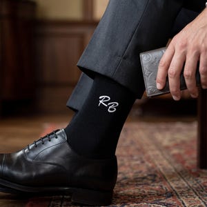 May include: A pair of black socks featuring white embroidered initials RB in a cursive script, worn with dark grey trousers and a black leather dress shoe. The socks are solid black and reach mid-calf, showcasing the white monogram detail clearly.