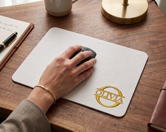 Op de afbeelding: Een rechthoekige witte muismat met een goudkleurig cirkelvormig embleem met de naam OLIVIA in het midden gedrukt. De muismat heeft afgeronde hoeken en ligt op een houten bureauoppervlak met een zwarte computermuis erop.