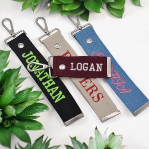 Pode incluir: Quatro chaveiros personalizados com ferragens prateadas. Os chaveiros são pretos, bege, bordô e azuis. O chaveiro preto tem o nome "JONATHAN" em verde. O chaveiro bege tem o nome "PETERS" em vermelho. O chaveiro bordô tem o nome "LOGAN" em branco. O chaveiro azul tem o nome "SARA" em rosa.