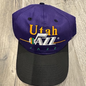 May include: A purple and black baseball cap with the words "Utah Jazz" embroidered in gold and white. The cap has a black peak and a button on top. The team's logo is also embroidered on the front.