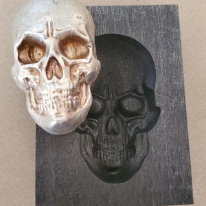 May include: A silver skull casting mold with a silver skull casting inside. The mold is made of black rubber and has a textured surface.
