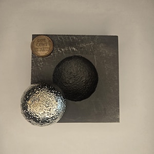 May include: A silver sphere with a textured surface sits on a black slate square. A copper coin with the inscription "ONE CENT UNITED STATES OF AMERICA" is visible in the top left corner.