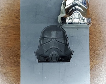 May include: A gray metal mold with a raised impression of a black Stormtrooper helmet. A silver Stormtrooper helmet is sitting on top of the mold.