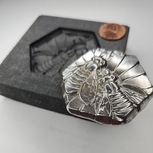 May include: A silver hexagonal mold with a detailed bee design. The mold is sitting on a gray stone surface.