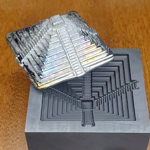 May include: A silver-colored pyramid-shaped mold with a matching pyramid-shaped casting resting on top. The mold is made of a dark gray material and has a textured surface. The casting is iridescent and reflects light.