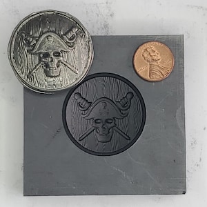 May include: A black metal mold with a pirate skull design. The mold is on a gray surface next to a copper penny.