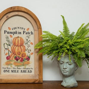 Può includere: Una stampa incorniciata con una cornice in legno rustica e uno sfondo in juta. La stampa presenta una pila di zucche, mais, mele e girasoli con il testo "Country Pumpkin Patch" e "One Mile Ahead".