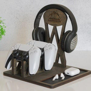 May include: A wooden stand with a personalised nameplate that says "HARRISON" and a mountain design. The stand holds a pair of black headphones, two white video game controllers, and a pair of white earbuds.