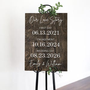 May include: A dark wooden sign with the words "Our Love Story" in white script. The sign displays significant dates: 06.13.2021, 10.16.2024, and 08.23.2026, along with the names "Emily & William." The sign is supported by a black metal stand and adorned with greenery.