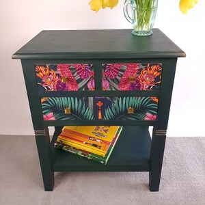 May include: A dark green wooden side table with two drawers and a lower shelf. The drawers are decorated with a colourful tropical print featuring dragons and palm leaves. The table has gold accents and is topped with a bouquet of yellow tulips in a blue glass jug.