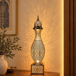 May include: A tall, metallic table lamp with an intricate, perforated pattern that casts a detailed shadow. The lamp features a bulbous body, a narrow neck, and a domed top, all resting on a square, illuminated base with matching cutouts.