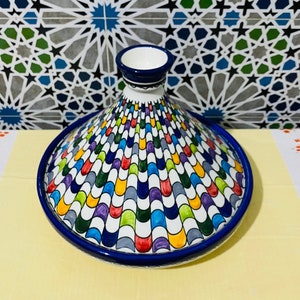 May include: A colourful ceramic tagine with a blue rim and a white base. The tagine has a geometric pattern of colourful, overlapping arches.
