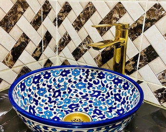 May include: A white ceramic sink with a blue and white floral pattern. The sink has a gold faucet and is set against a white and brown tiled wall.