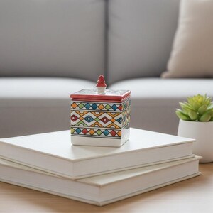May include: A decorative ceramic box with a geometric pattern in red, yellow, blue, and green sits atop two white books. The box has a red lid with a small, pointed finial. A small succulent in a white pot is also on the table.