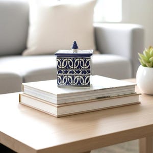 Puede incluir: Una caja decorativa cuadrada azul y blanca con tapa, con un patrón geométrico repetitivo. La caja está colocada encima de dos libros sobre una mesa de centro de madera. Un sofá gris y una pequeña planta en maceta están en el fondo.