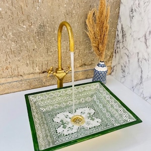 Puede incluir: Un lavabo cuadrado de cerámica verde y blanca con un patrón decorativo. Un grifo dorado vierte agua en el lavabo. Un jarrón con plantas secas se encuentra a la derecha del lavabo. El lavabo tiene un desagüe dorado.