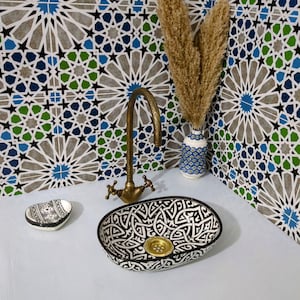 Pode incluir: Uma pia oval de cerâmica preta e branca com um ralo dourado, um pequeno saboneteira e uma torneira de latão vintage. O fundo apresenta uma parede de azulejos estampados com desenhos geométricos azuis, verdes e cinzentos. Há também um vaso com plantas secas.