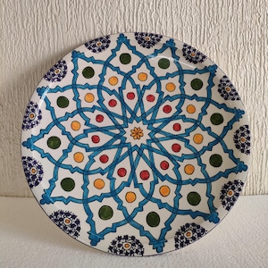 May include: A white ceramic plate with a blue, green, yellow, and red geometric pattern. The plate has a decorative border with small blue and purple flowers.