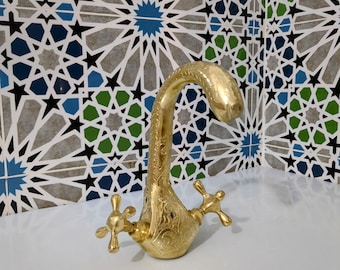 Op de afbeelding: Een goudkleurige kraan met een gebogen tuit en kruisvormige handgrepen. De kraan heeft een sierlijk, gegraveerd ontwerp. De achtergrond is een betegelde muur met blauwe, groene en zwarte geometrische vormen.