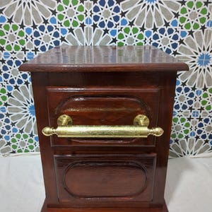 Puede incluir: Una mesita de noche de madera marrón oscuro con un tirador de latón. La mesita tiene forma rectangular con un diseño ovalado en la parte delantera. El fondo presenta un diseño de azulejos estampados en azul, verde y blanco.