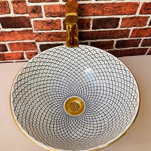May include: A white ceramic sink with a black and white geometric pattern and a gold rim. The sink has a gold drain and is mounted on a brick wall.
