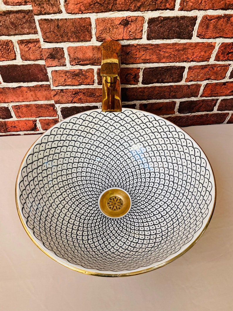 May include: A white ceramic sink with a black and white geometric pattern and a gold rim. The sink has a gold drain and is mounted on a brick wall.