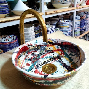 May include: A white ceramic sink with a colorful abstract design. The sink has a gold-colored drain and is sitting on a white surface. A gold-colored faucet is in the background.