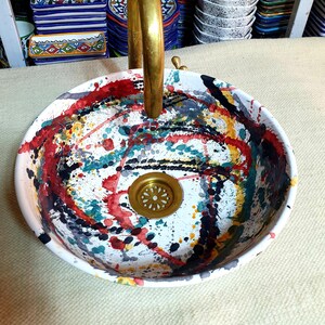 May include: A white ceramic sink with a brass faucet and a black, red, yellow, and green abstract design. The sink has a brass drain.