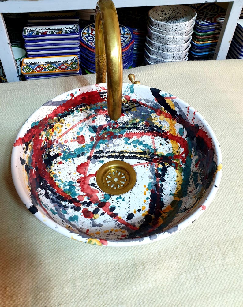 May include: A white ceramic sink with a brass faucet and a black, red, yellow, and green abstract design. The sink has a brass drain.