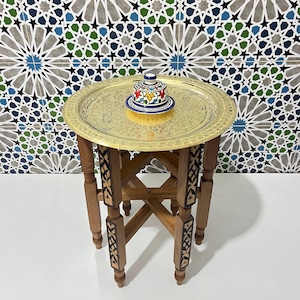 Può includere: Un tavolino rotondo con vassoio in ottone e base in legno con intarsi geometrici. Il tavolo ha quattro gambe collegate da una crociera. Un piccolo contenitore in ceramica con coperchio e motivi floreali dipinti a mano poggia sul vassoio.