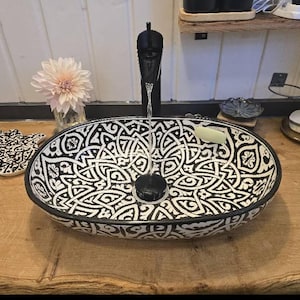 Puede incluir: Un lavabo ovalado de cerámica con un patrón geométrico en blanco y negro. Un grifo negro está encima del lavabo, con agua fluyendo. Una flor y un jabonera están sobre la encimera de madera. El lavabo es un accesorio de baño.