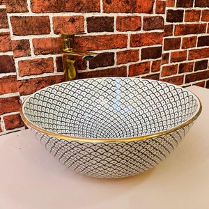 May include: White ceramic sink with a black and white geometric pattern and a gold rim. The sink is set against a brick wall with a gold faucet.