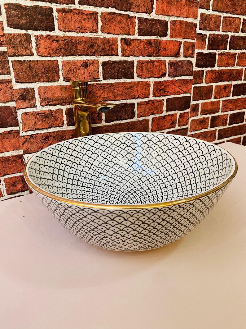 May include: White ceramic sink with a black and white geometric pattern and a gold rim. The sink is set against a brick wall with a gold faucet.