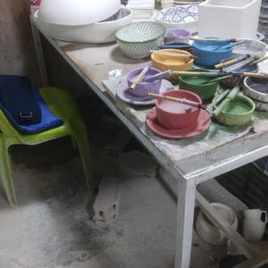 May include: A white table with a variety of colorful ceramic bowls and plates. Some of the bowls are filled with water and have paintbrushes in them. There are also some wooden sticks in the bowls.