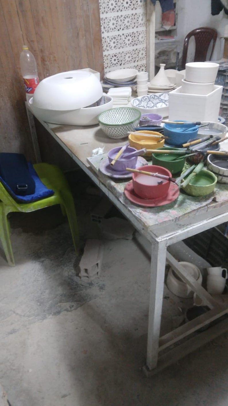 May include: A white table with a variety of colorful ceramic bowls and plates. Some of the bowls are filled with water and have paintbrushes in them. There are also some wooden sticks in the bowls.