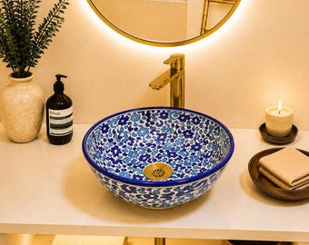 Pode incluir: Uma cuba de apoio redonda em cerâmica com um padrão floral azul e branco vibrante no interior e exterior. A cuba inclui uma válvula de escoamento em metal dourado e uma torneira dourada a condizer, montada sobre uma bancada branca.