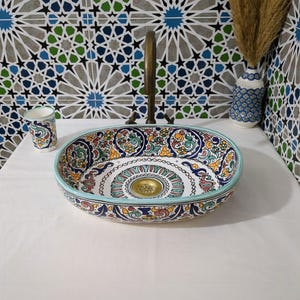 Könnte beinhalten: Ein ovales Keramikwaschbecken mit einem farbenfrohen Blumenmuster und einem hellblauen Rand. Ein passender Becher und eine Vase mit blauem Blumendesign befinden sich auf der weißen Oberfläche. Über dem Waschbecken ist ein Wasserhahn aus Messing montiert, im Hintergrund eine gemusterte Fliesenwand.