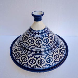 Puede incluir: Una olla de tagine de cerámica azul y blanca con tapa cónica. La tapa y la base presentan intrincados patrones geométricos en azul oscuro sobre un fondo blanco. El tagine es un recipiente de cocina tradicional del norte de África.