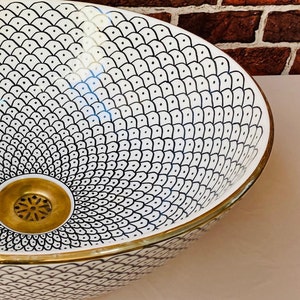 May include: A white ceramic sink with a black and white geometric pattern. The sink has a gold rim and a gold drain.