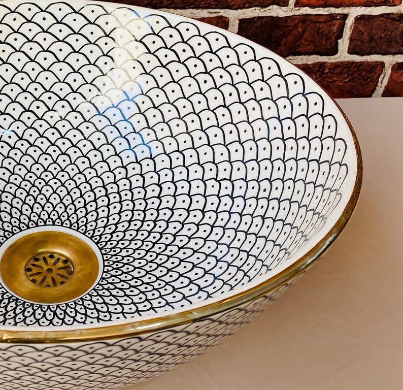 May include: A white ceramic sink with a black and white geometric pattern. The sink has a gold rim and a gold drain.