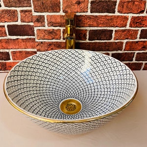 May include: A white ceramic bathroom sink with a black and white geometric pattern and a gold rim. The sink has a gold drain.