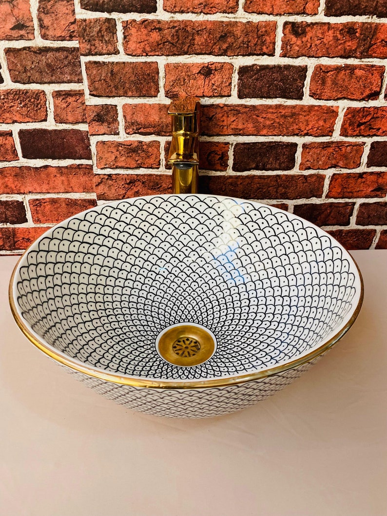 May include: A white ceramic bathroom sink with a black and white geometric pattern and a gold rim. The sink has a gold drain.