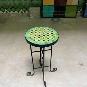 Peut inclure: Une table d'appoint ronde dotée d'un plateau en mosaïque au motif géométrique dans des tons de vert, jaune et bleu. La table est soutenue par une structure en métal noir avec quatre pieds fins incurvés à la base et une entretoise centrale.