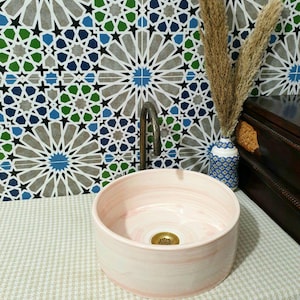 May include: A pink ceramic vessel sink with a white swirl pattern. The sink is round and has a drain in the centre. The sink is sitting on a white and grey patterned surface.