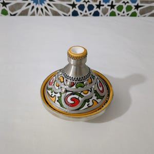 May include: A silver-colored tagine pot with a conical lid and a wide, shallow base. The base is decorated with a colorful, intricate pattern of red, yellow, green, and white designs. The lid has a decorative rim.