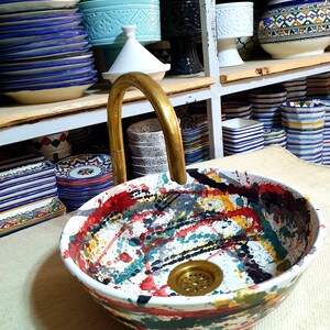 May include: A white ceramic sink with a colorful abstract design. The sink has a brass drain and a gold faucet. The sink is on a white countertop.