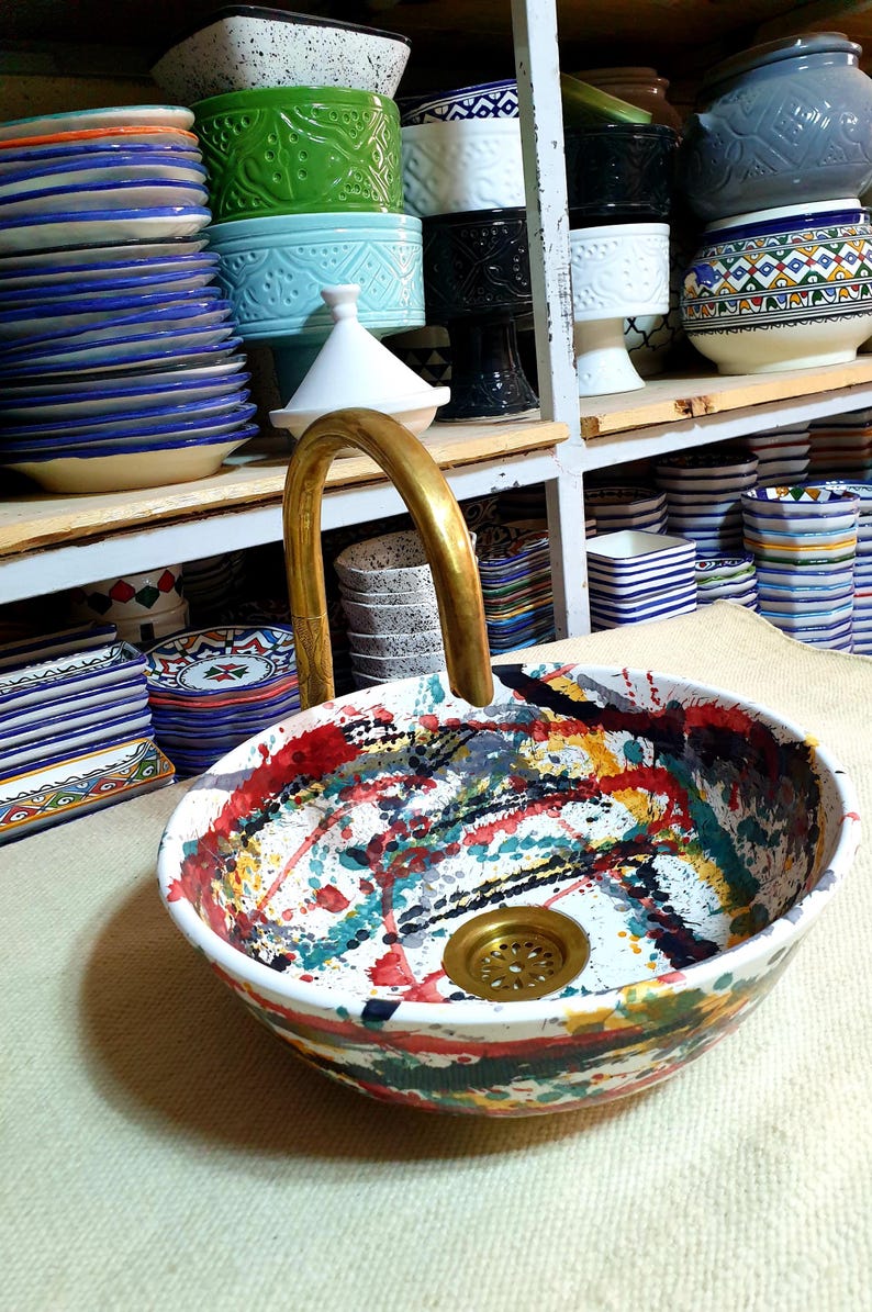 May include: A white ceramic sink with a colorful abstract design. The sink has a brass drain and a gold faucet. The sink is on a white countertop.