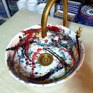 May include: A white ceramic sink with a gold faucet and a colorful abstract design. The sink has a drain in the center and is decorated with splashes of red, blue, green, yellow, and black paint.